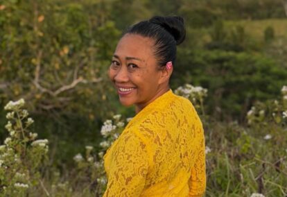 Smiling woman with dark hair in a bun wearing a yellow lace top standing outdoors among wildflowers and greenery